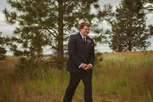 Groom in navy suit standing thoughtfully in field with pine trees on overcast day.