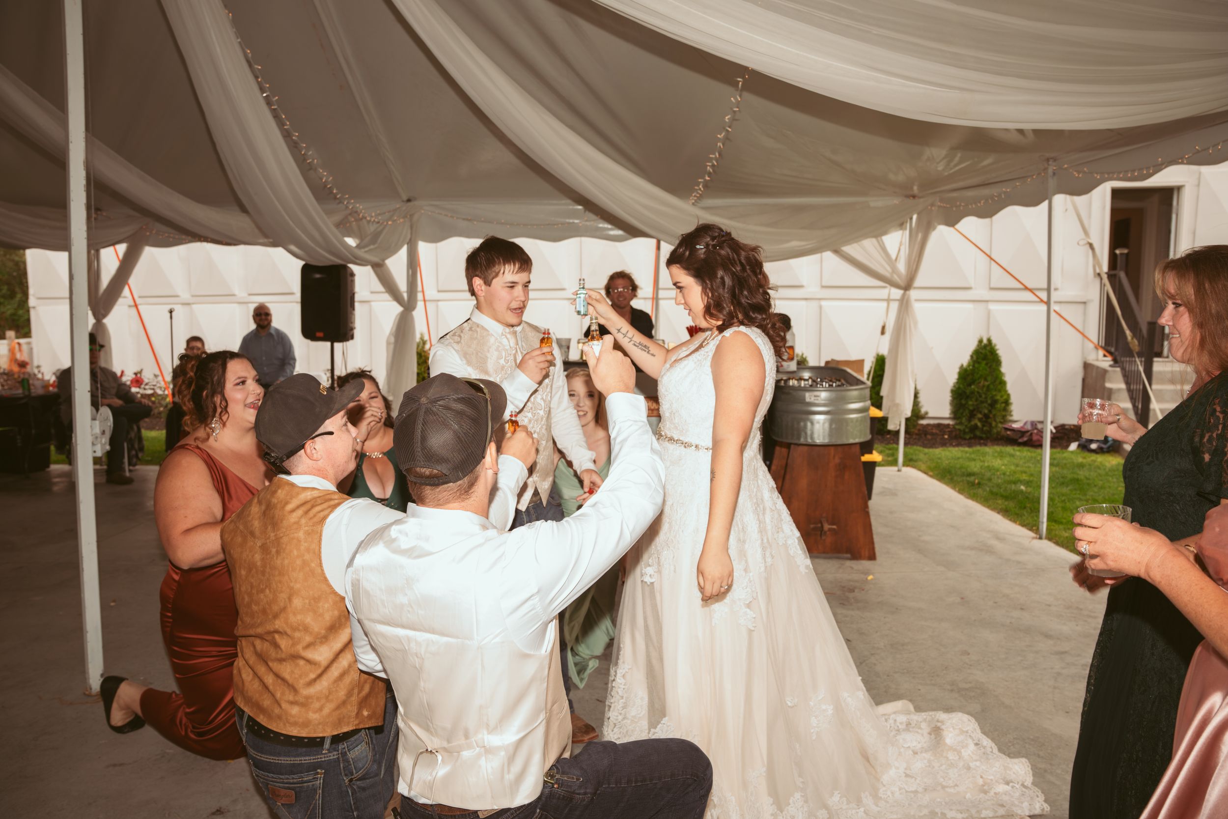 Wedding guests dancing and celebrating together under a white tent draped with string lights at an outdoor reception.