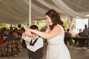 A bride in a white dress dances joyfully with a young guest under an outdoor wedding tent.
