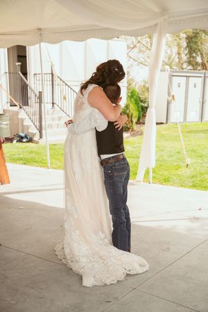 A bride and groom share an intimate embrace under a white tent at an outdoor wedding ceremony.