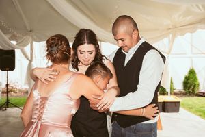 A group embraces in a tight emotional hug during a wedding reception under a white tent.
