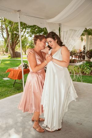 Two people in elegant dresses share an intimate moment under a white tent canopy at an outdoor event.