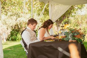 A couple seated at an elegant outdoor table with a black tablecloth enjoys a meal under a white canopy.