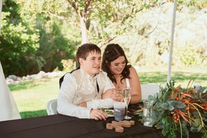 Wedding party seated at a decorated head table with greenery under a white tent.
