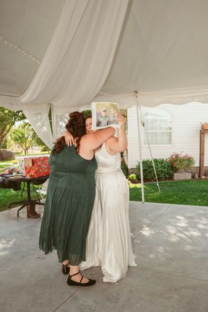 A wedding guest in a green dress embraces someone in a white wedding gown under an outdoor tent reception.