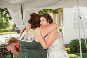 Two people share an emotional embrace under a white tent at an outdoor wedding reception.