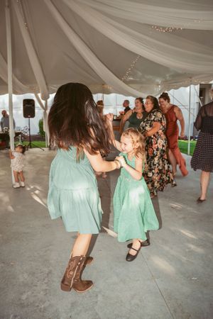 Dancers in matching mint green dresses twirl together on a concrete floor under a draped tent at a celebration.