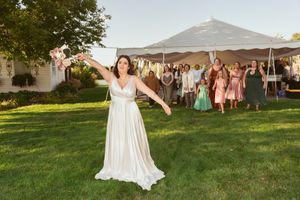 A bride in a white gown poses joyfully on a lawn with wedding guests in colorful dresses in the background.