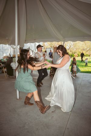Dancers at a wedding reception having fun under a white tent, with one wearing cowboy boots and a mint dress.
