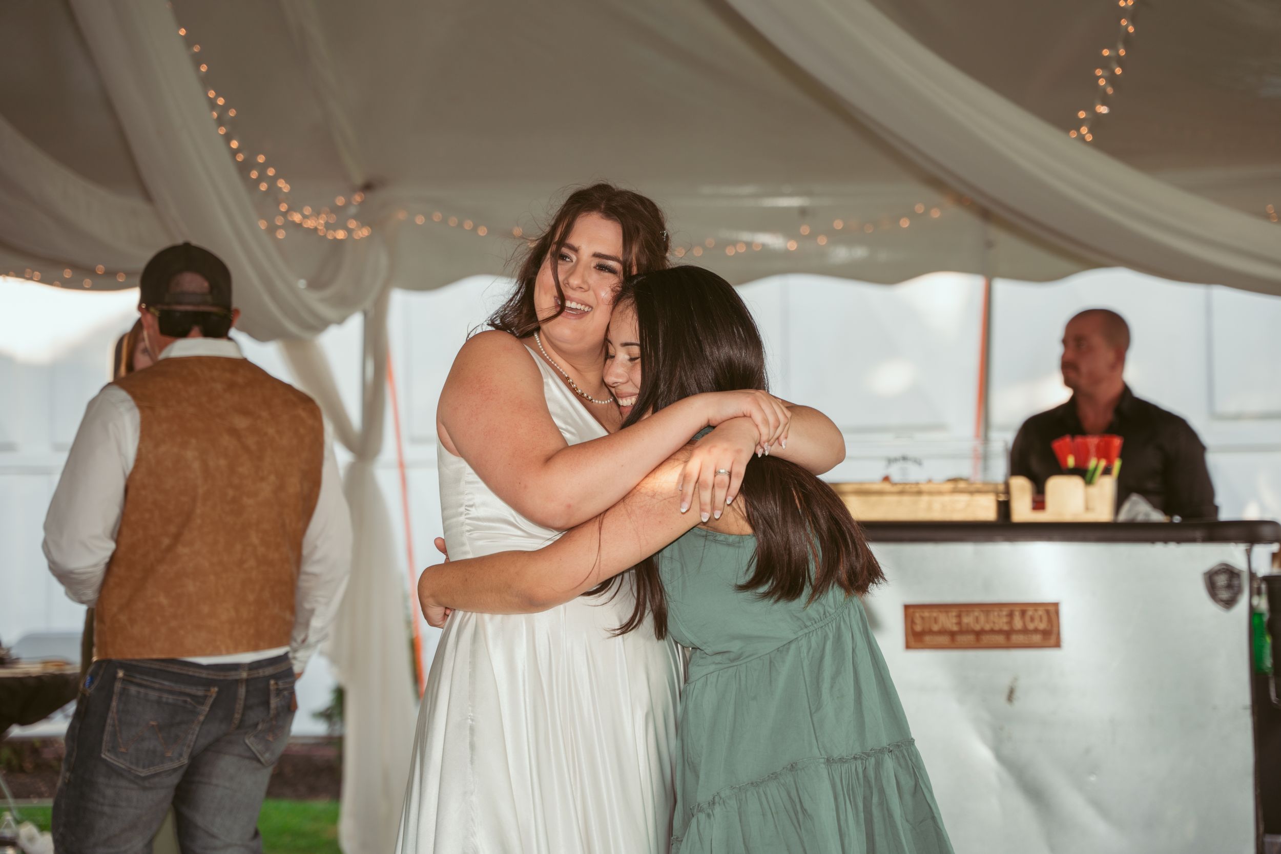 Emotional embrace between people at a wedding reception with fairy lights and decor visible.