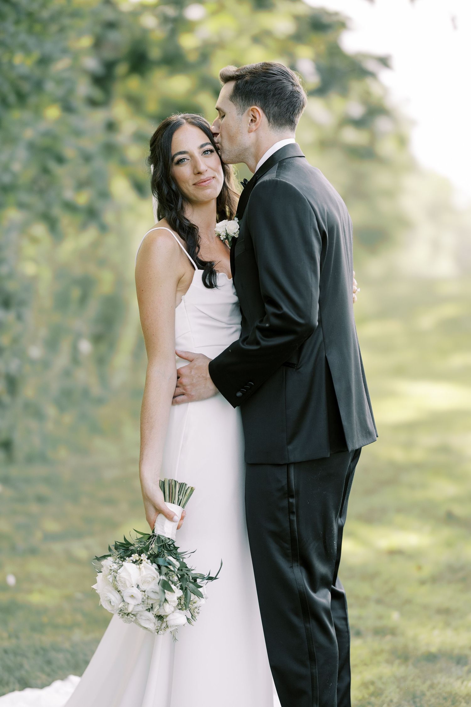 A tender wedding moment captured outdoors with a beautiful white bouquet and elegant formal attire.
