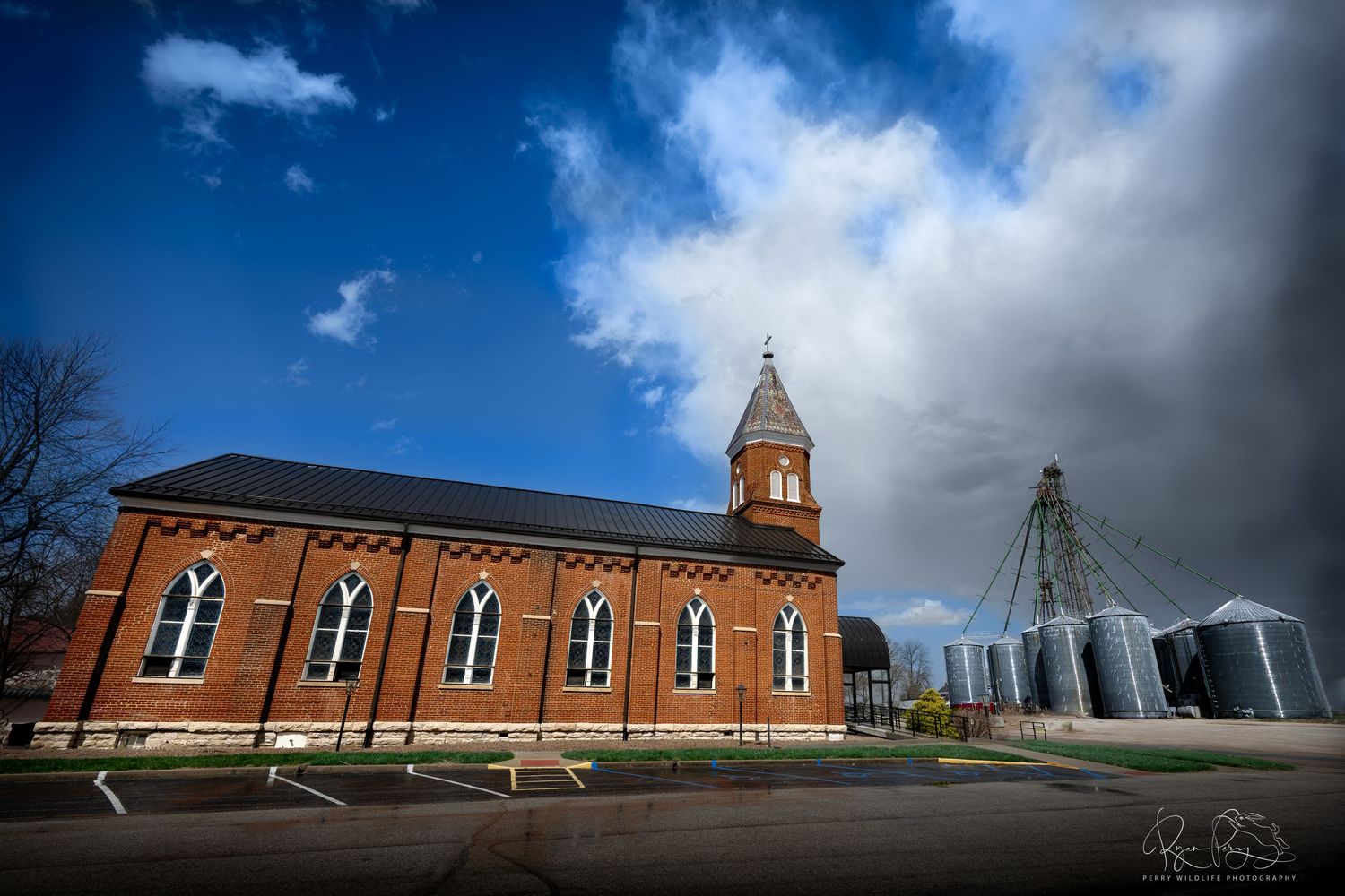 Red brick church with tall spire next to modern grain silos under dramatic clouds.