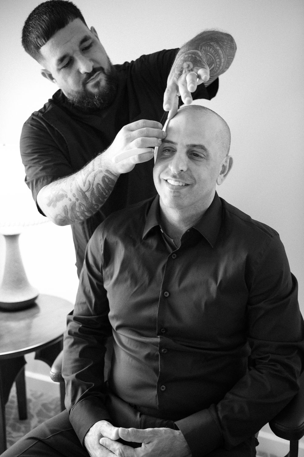A barber with tattoos working on a client's head in a professional black and white portrait.