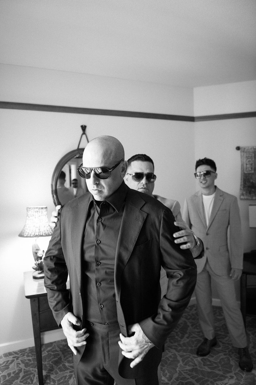 A black and white photo showing three people in sunglasses and formal attire posing dramatically indoors.