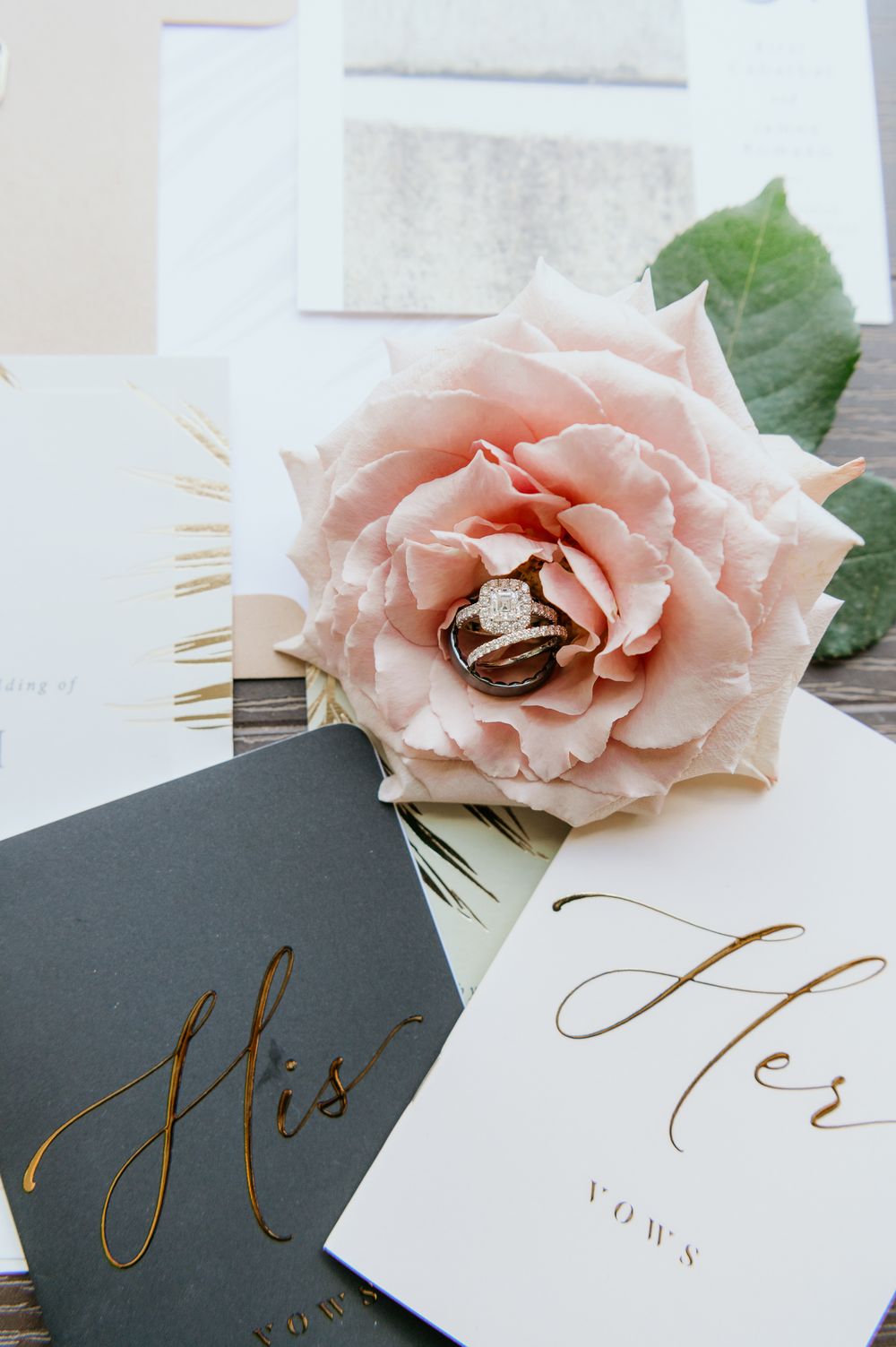 A pink rose rests on wedding vow cards with elegant gold 'His' and 'Vows' calligraphy text.