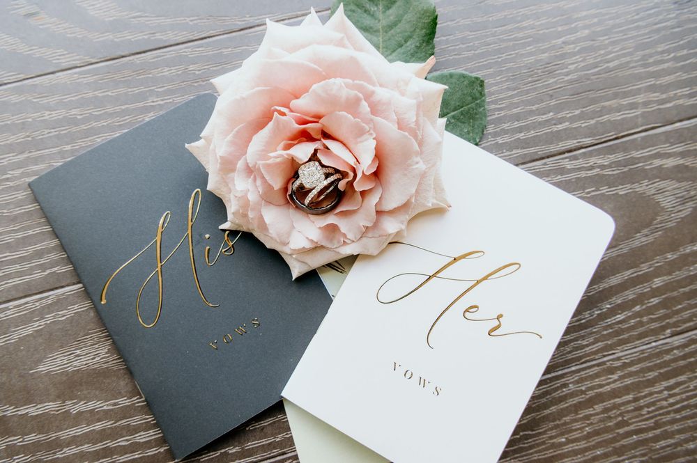 Wedding vows books in white and charcoal gray with gold lettering and a pink rose on wooden table.
