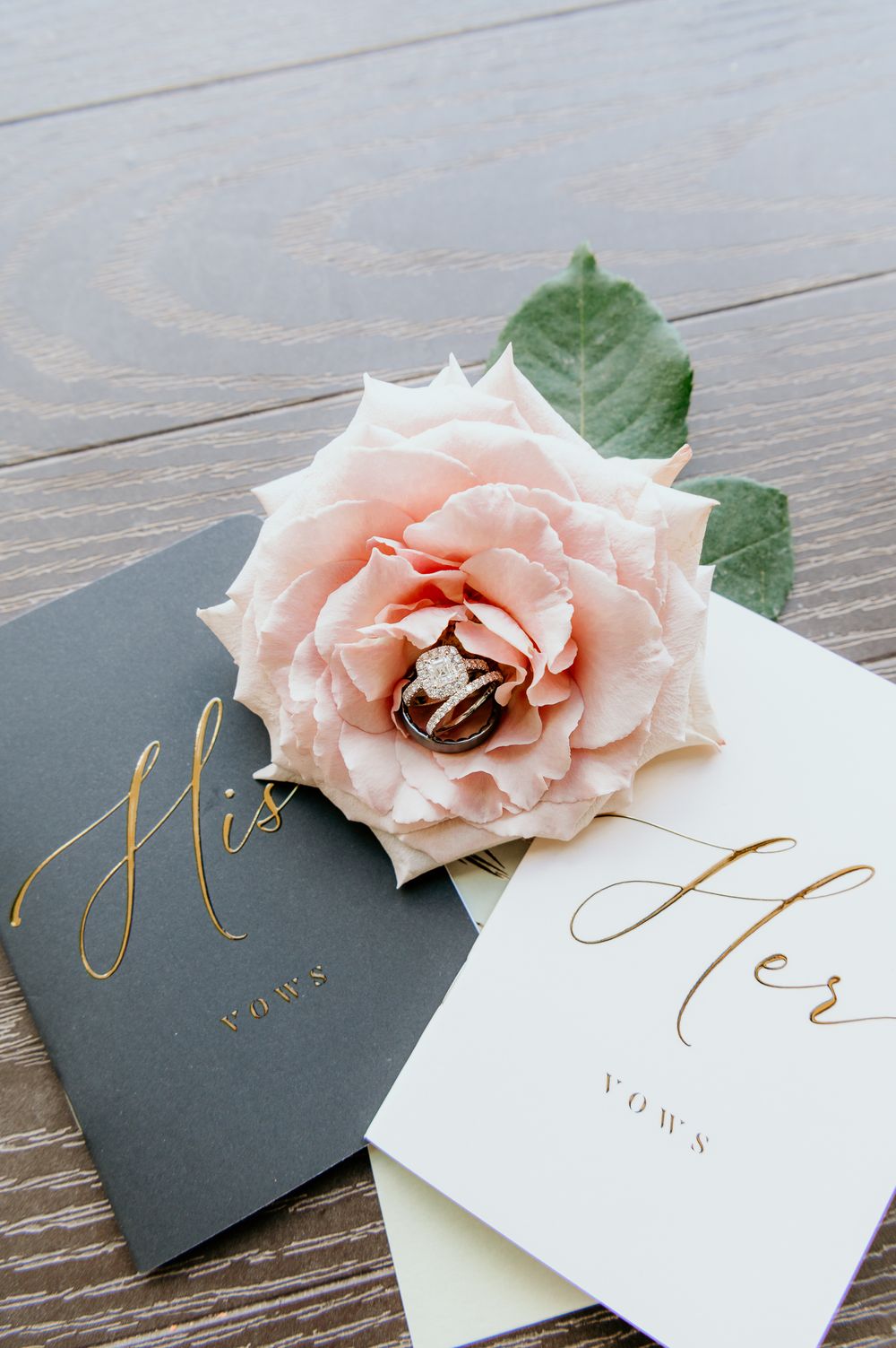 Wedding vow books in gray and white with gold lettering placed beside a delicate pink rose on wooden surface.