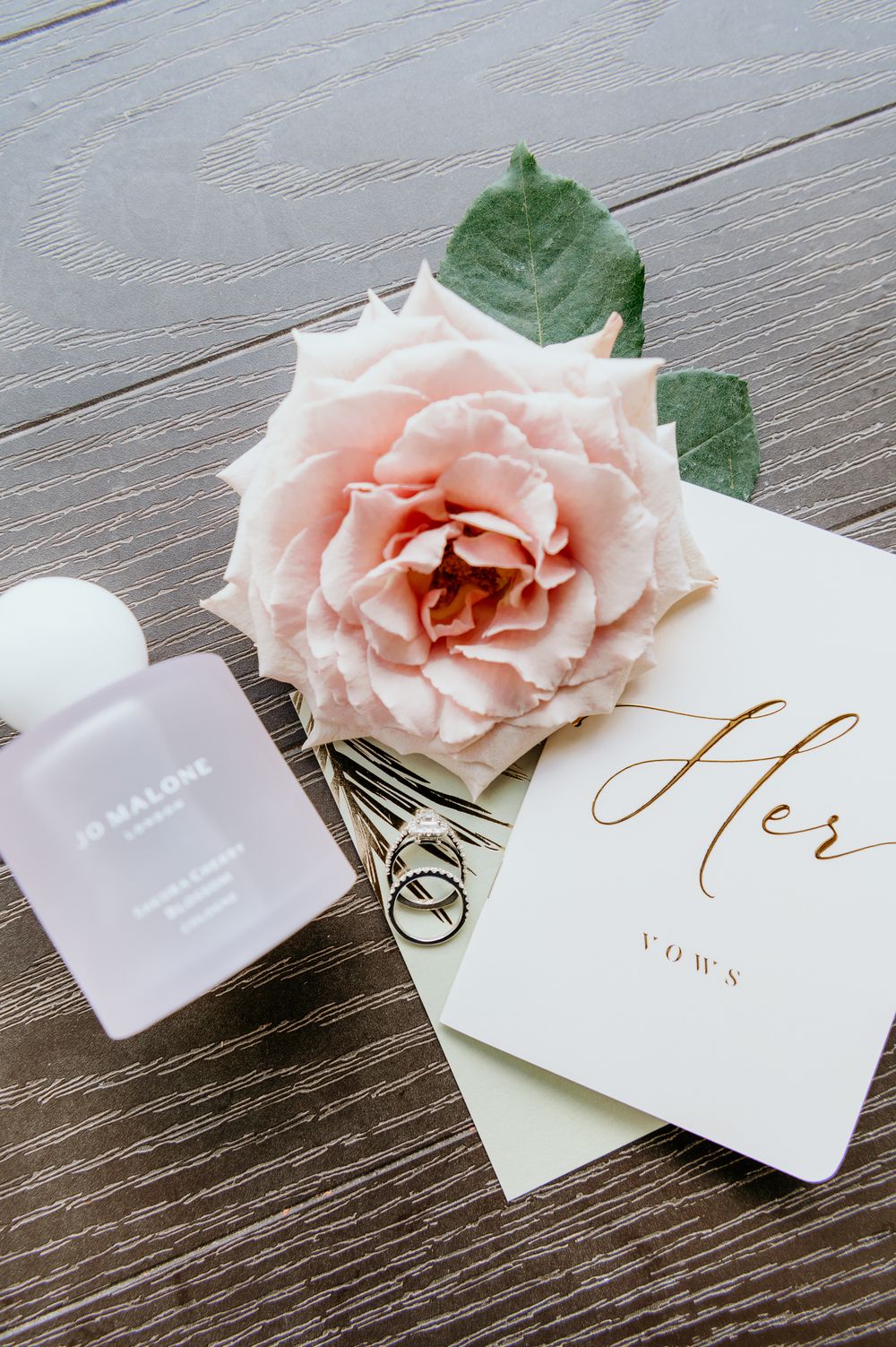 A blush pink rose rests beside a wedding vow book and perfume bottle on a rustic wooden surface.
