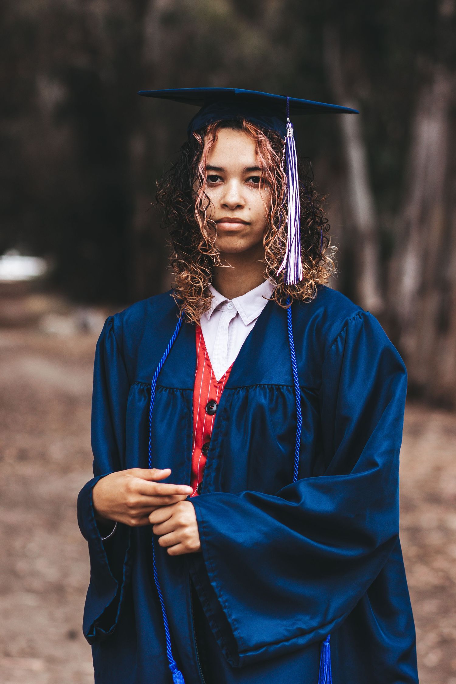 Graduate in royal blue cap and gown poses in wooded setting.