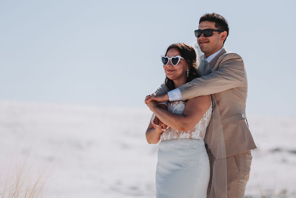 A couple wearing sunglasses embrace on a beach, the bride in white dress and groom in tan suit.