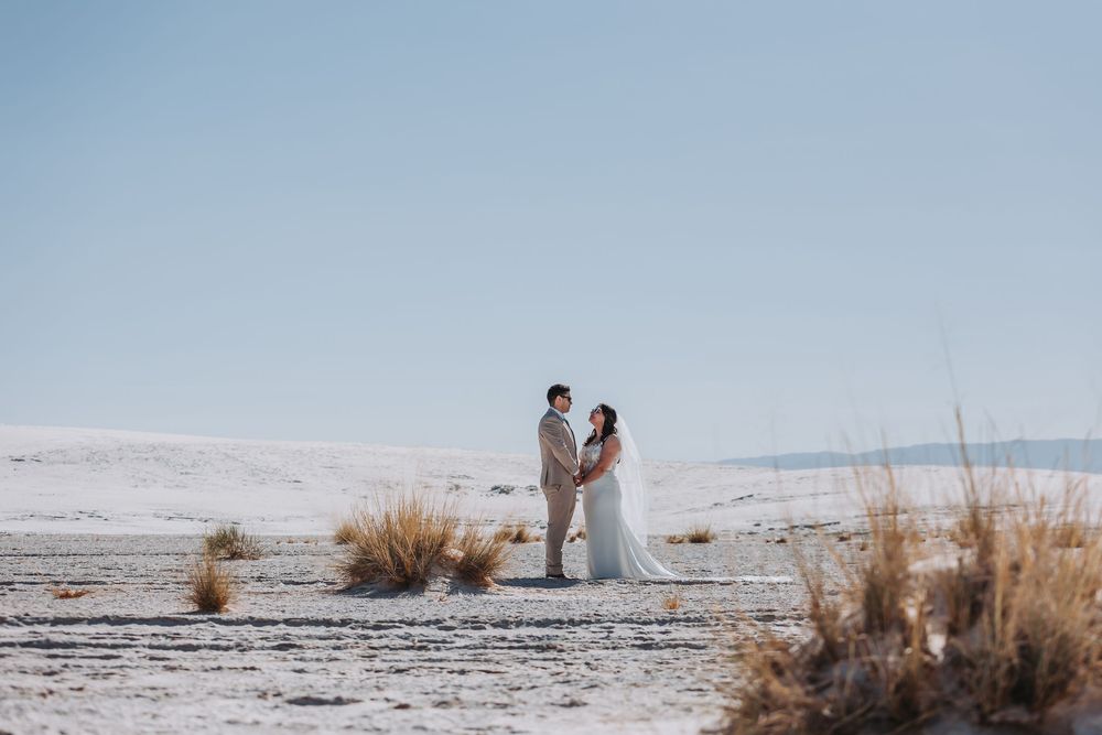 A couple in wedding attire stands together on snowy white sand dunes during golden hour.