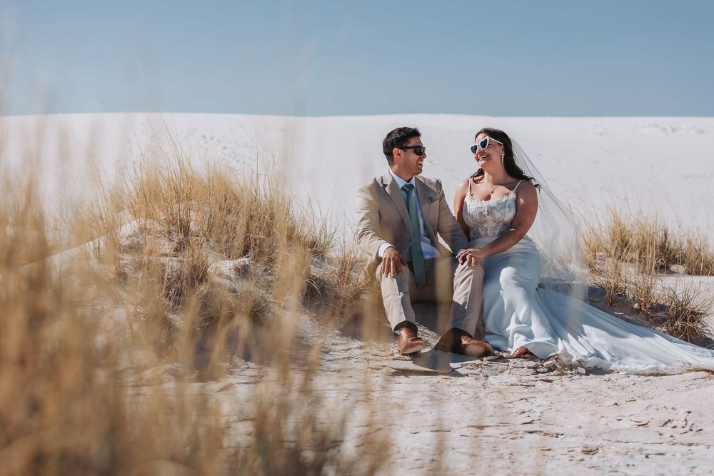 A couple sits together romantically on a tranquil beach amidst sand dunes and sea grass during golden hour.