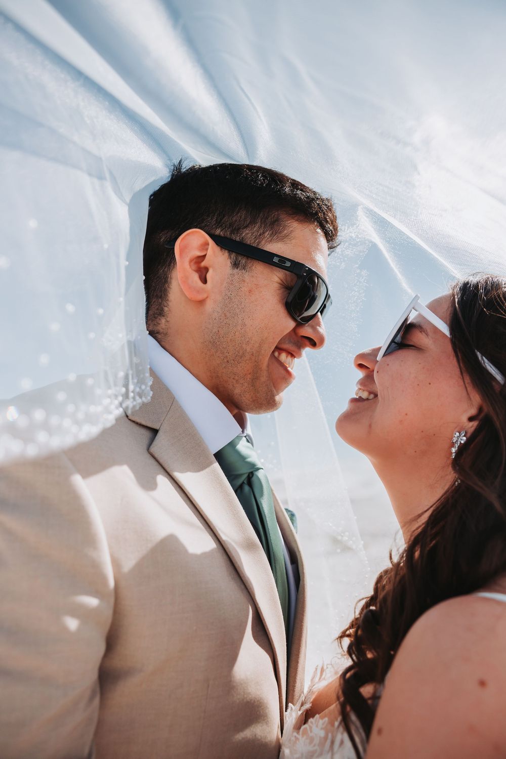 Joyful romantic moment between newlyweds under a flowing white veil against bright sky.