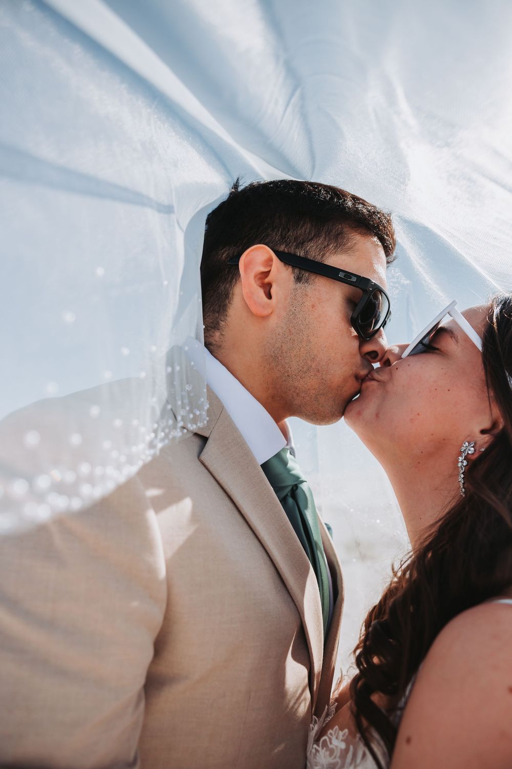 Romantic wedding photo showing couple kissing under flowing white veil against bright sky.