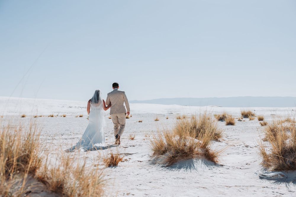 Newlyweds walking hand in hand on white sandy beach with desert brush and mountains in background.