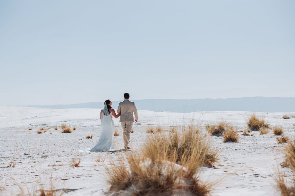Newlywed couple walking hand in hand across white sand dunes with mountains in background.