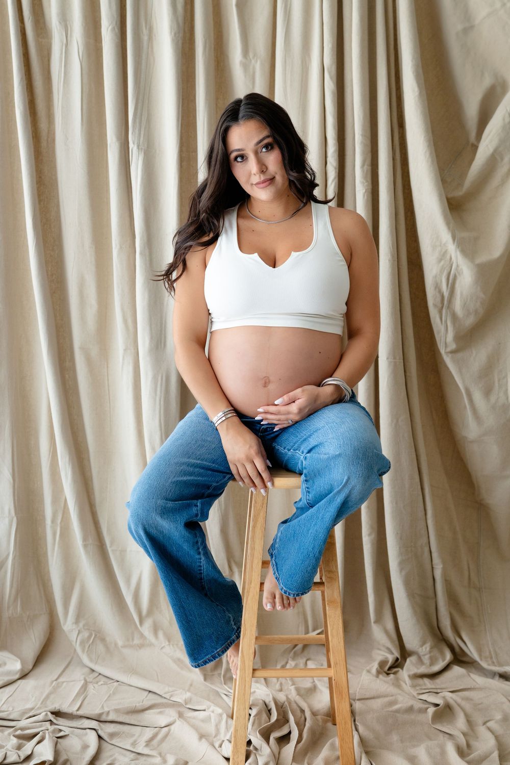Expectant woman in white crop top and jeans sits on wooden stool against beige curtain backdrop during maternity shoot.