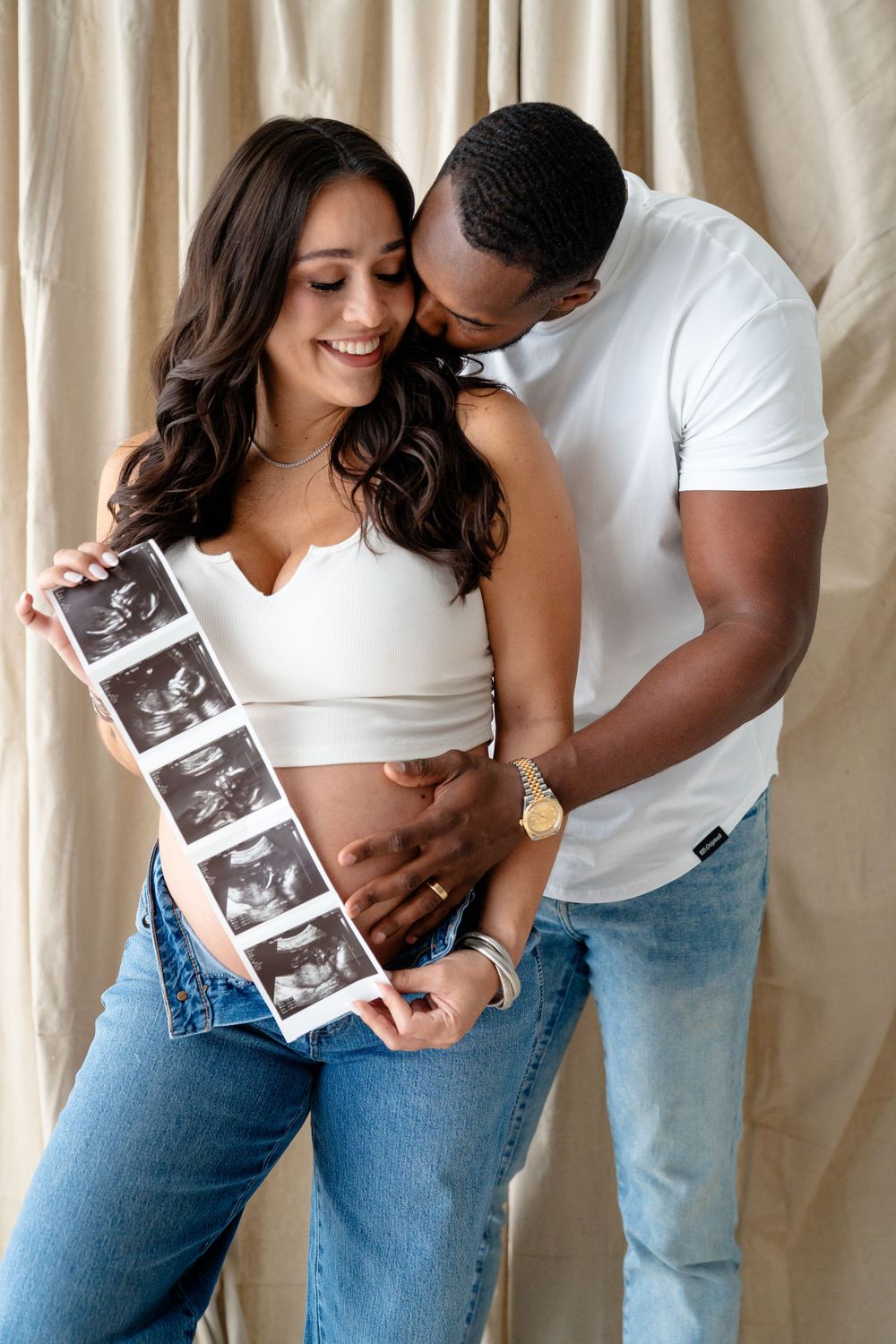 A couple shares an intimate moment while looking at ultrasound photos against a white curtain background.