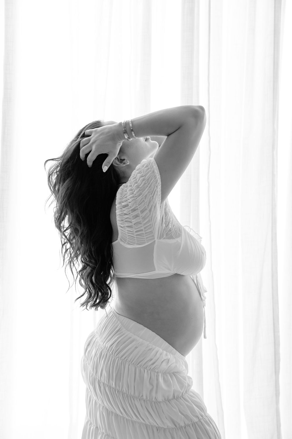 Black and white silhouette of an expectant mother in white lace maternity wear posing against a backlit window.