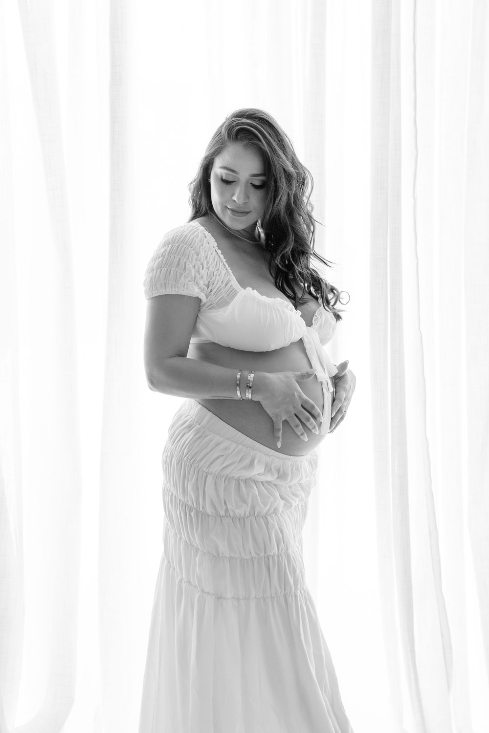 Three artistic black and white maternity photos show an expectant mother in a flowing white dress by sheer curtains.