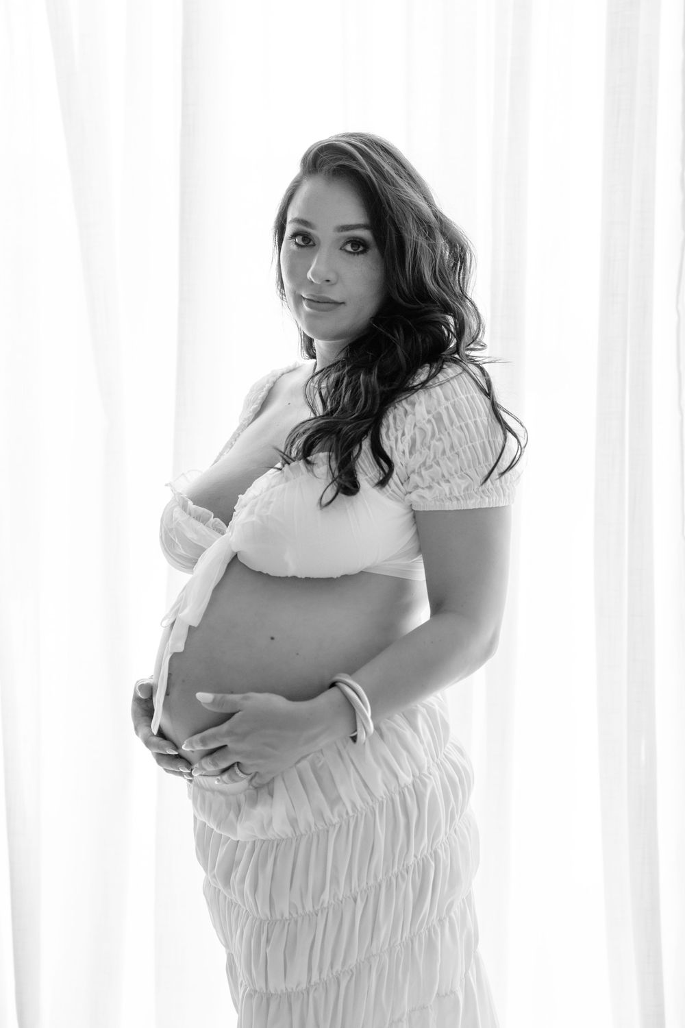 Black and white maternity photo of an expectant mother in a white dress standing by a window with natural light.