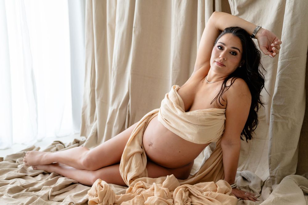 Expecting person rests on beige bedding by window with flowing curtains in soft natural lighting.