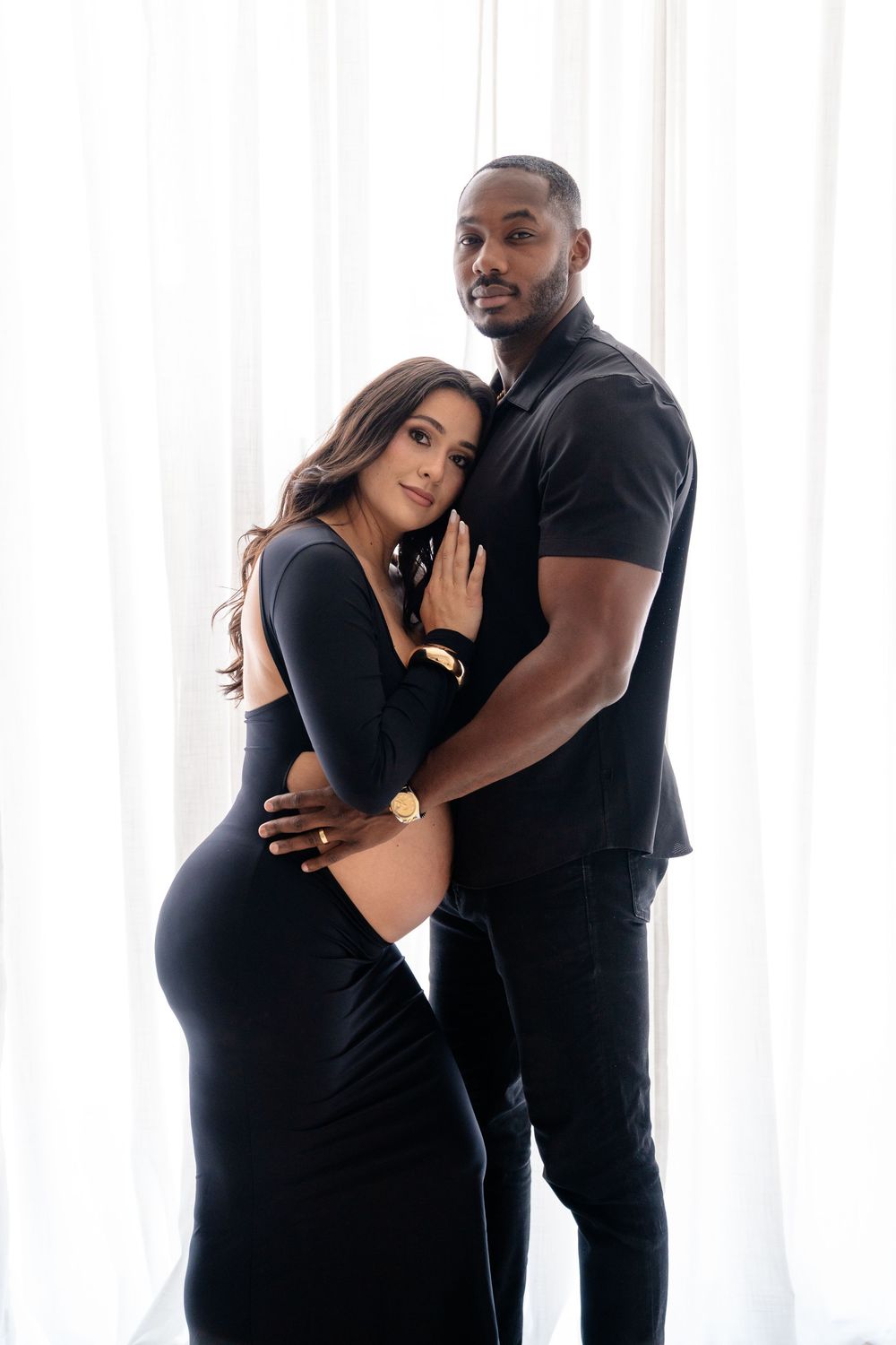 Elegant maternity photo of an expecting couple in matching black outfits embracing in front of white curtains.