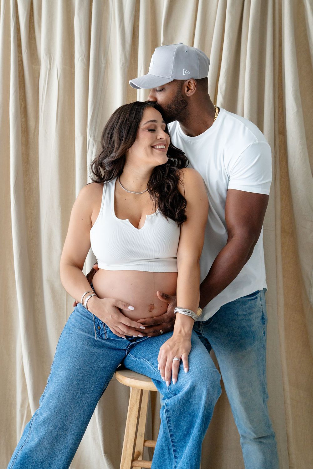 Intimate interracial couple sharing a tender moment against beige curtains, wearing casual white and denim outfits.