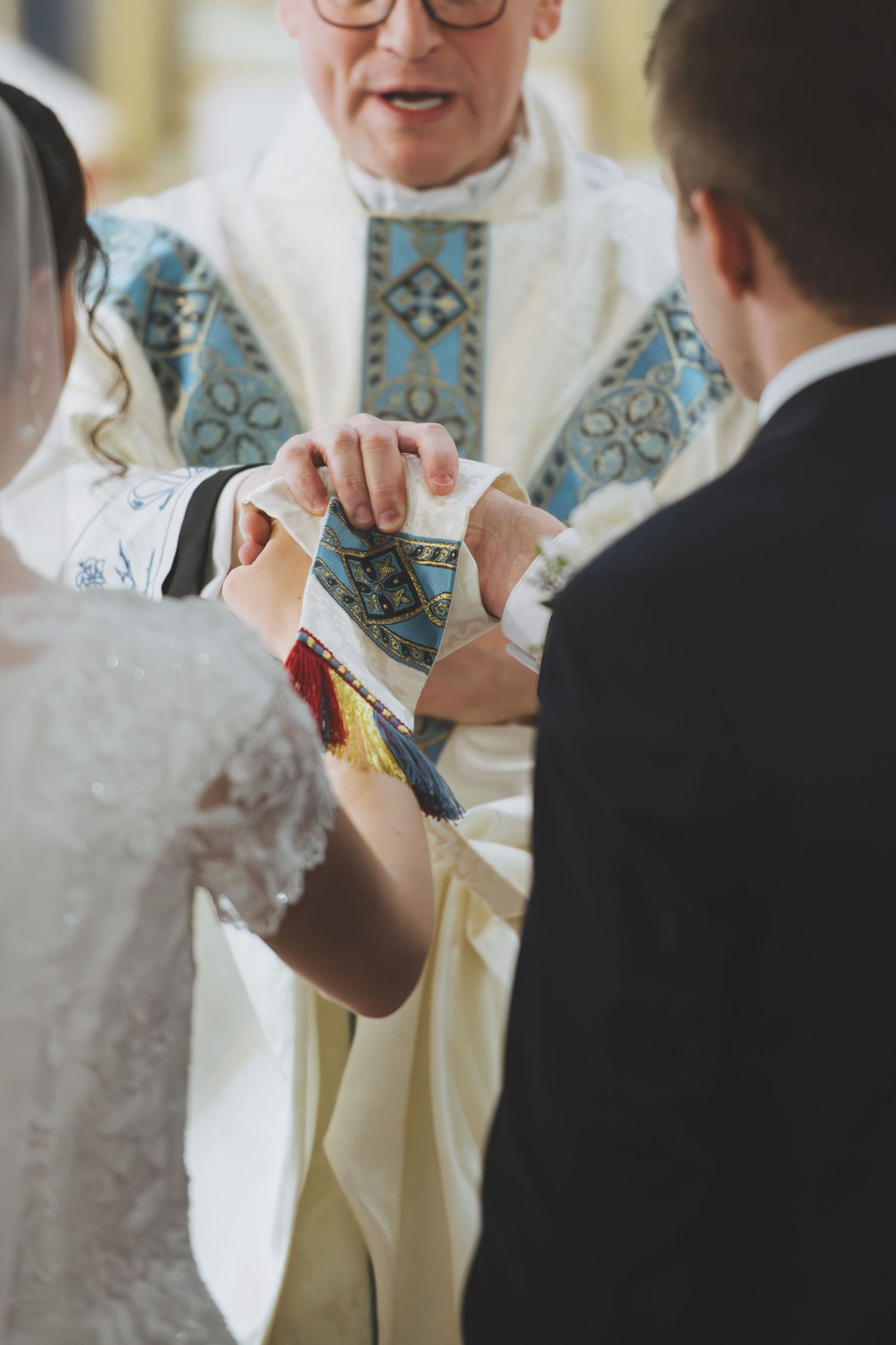 A priest in blue and white vestments performs a wedding ceremony blessing as couple's hands join together.
