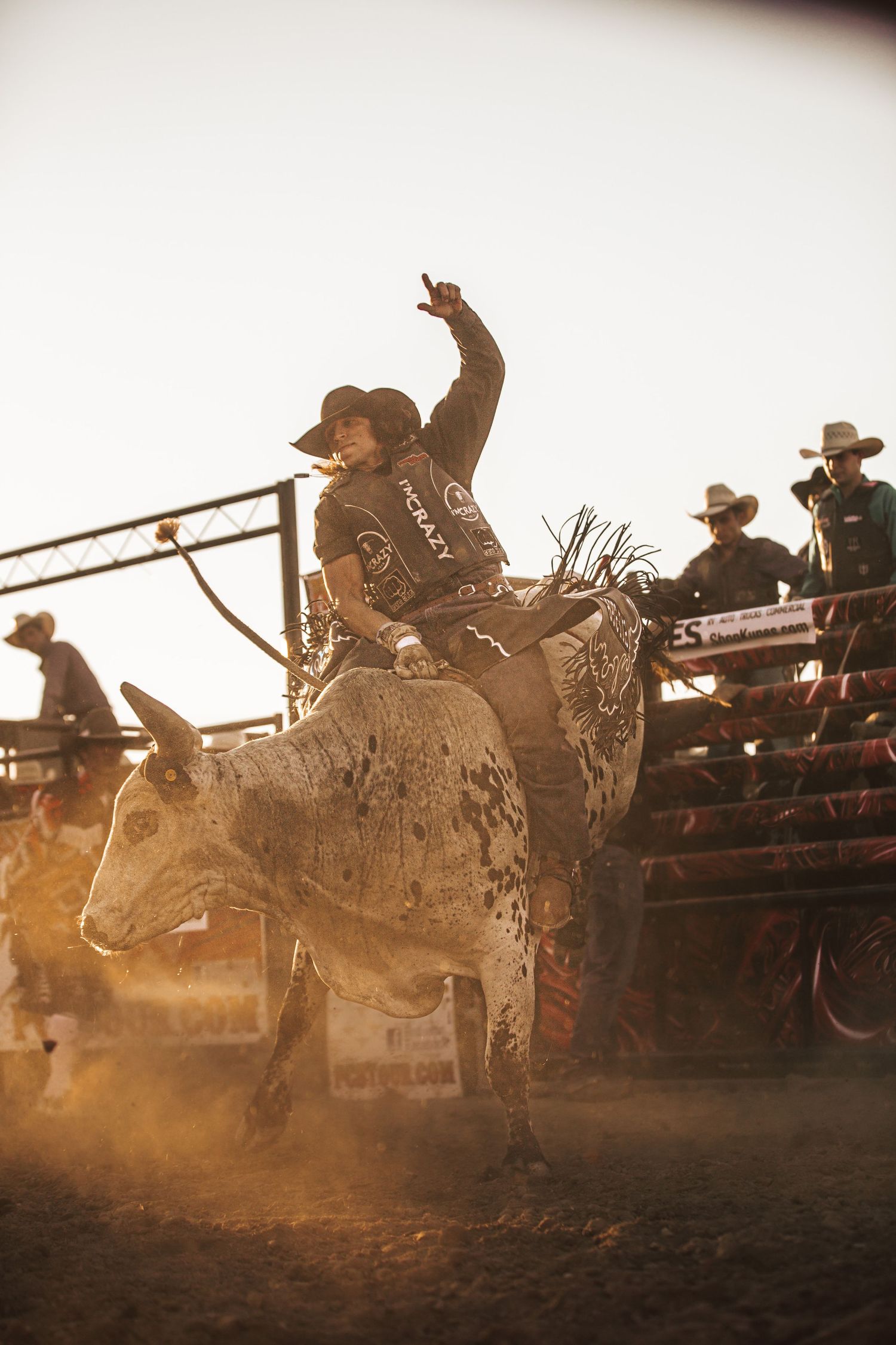 A rodeo cowboy rides a bucking bull in an arena during a sunset competition with spectators looking on from stands.