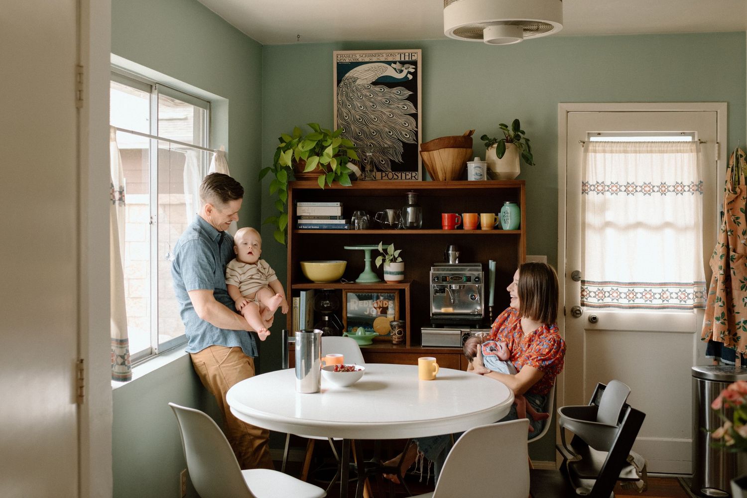 A green dining room features a modern round dining table with contrasting dark shelving and white trimmed windows.