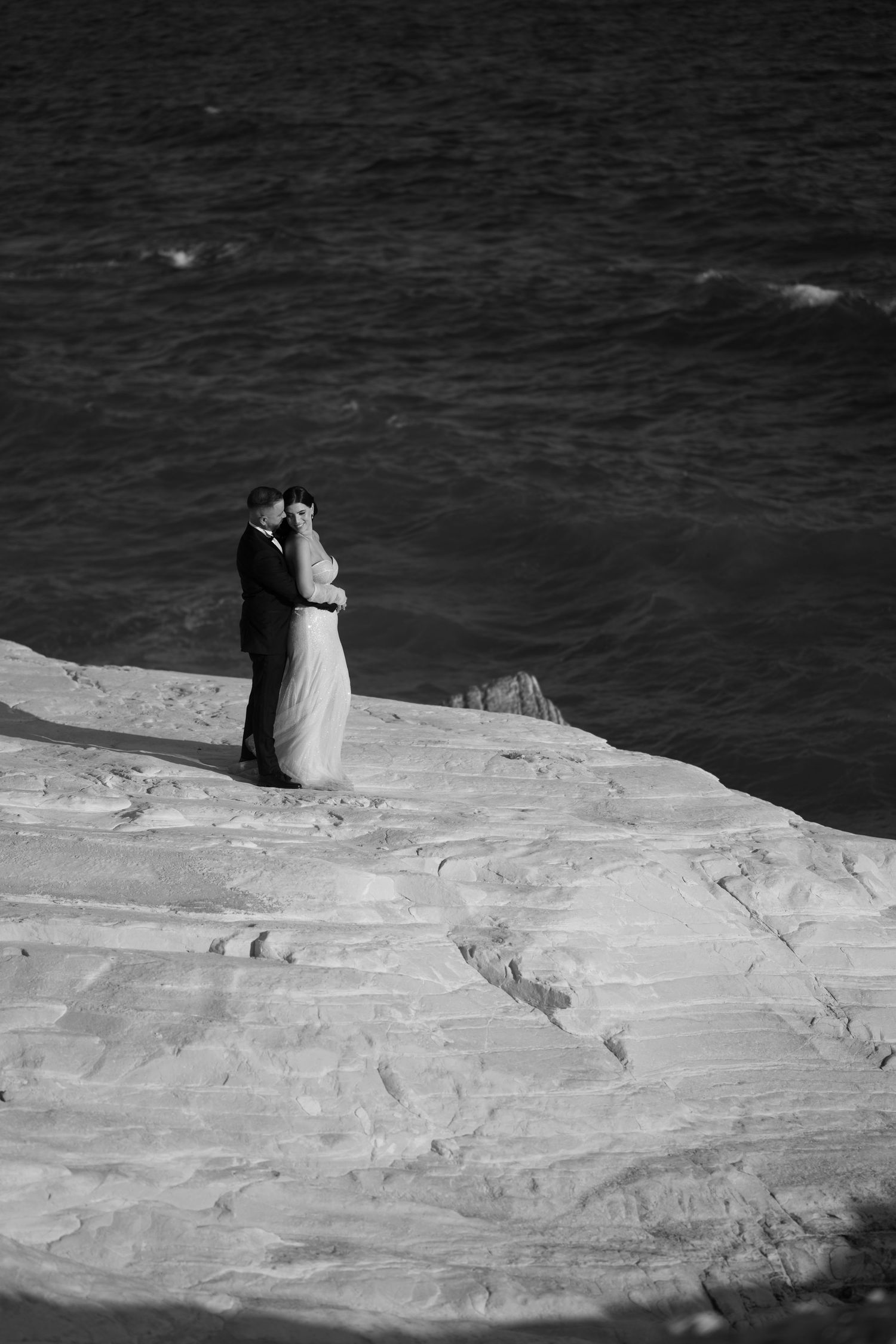 A romantic couple embrace on a dramatic white cliff overlooking dark ocean waters in black and white photography.