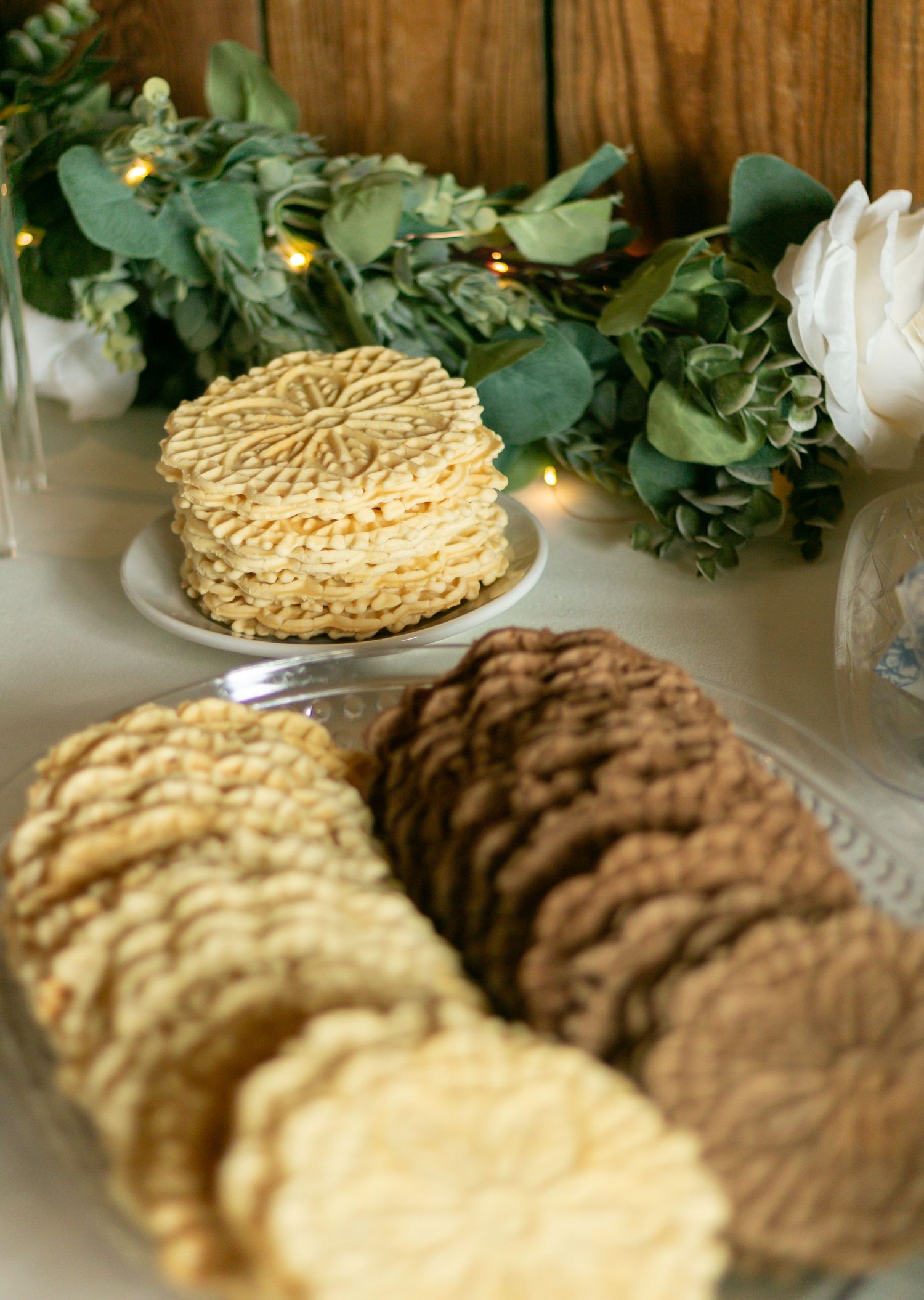 Stacks of golden pizzelle cookies and chocolate cookies arranged on plates with greenery and white flowers in the background.