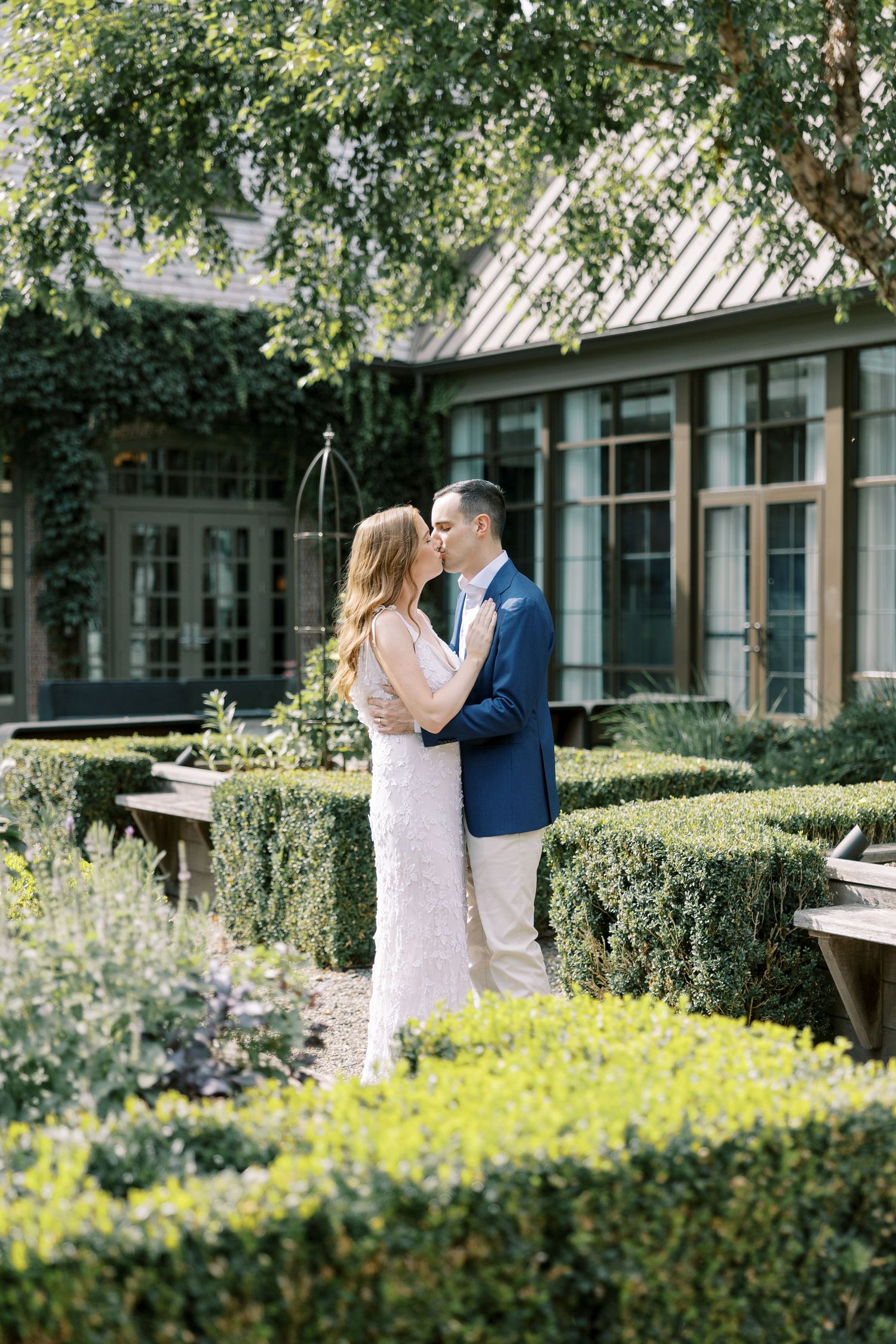 A couple shares an intimate moment in a garden setting with manicured hedges and flowering yellow plants.