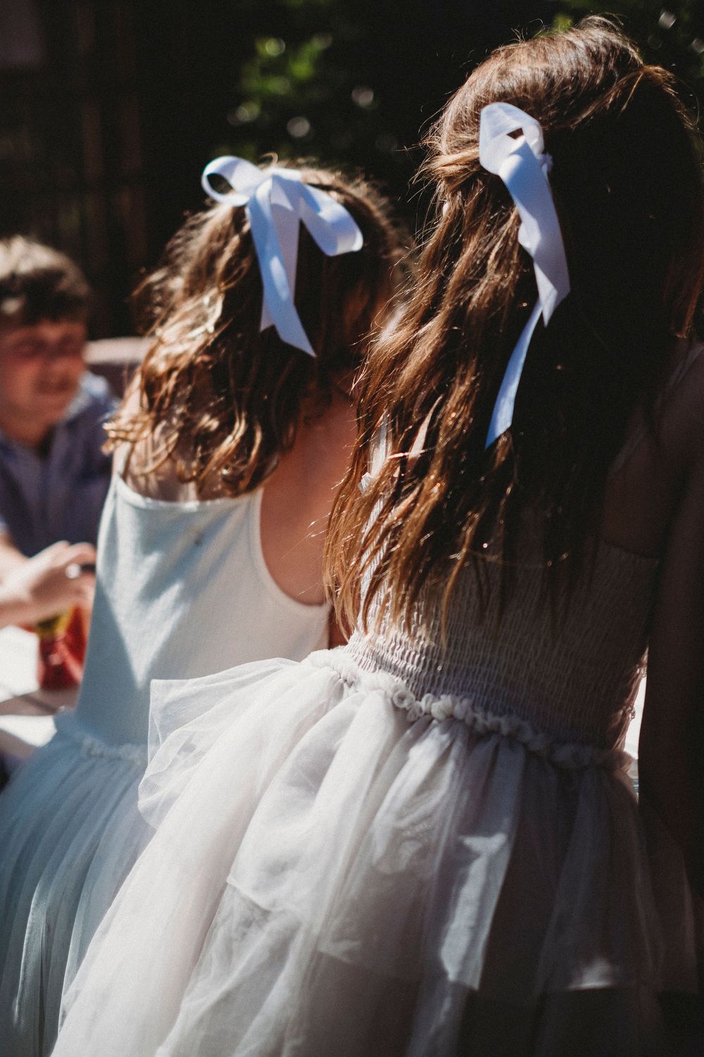 Two flower girls with white ribbon bows in their brown hair wearing white tulle dresses at an outdoor wedding.