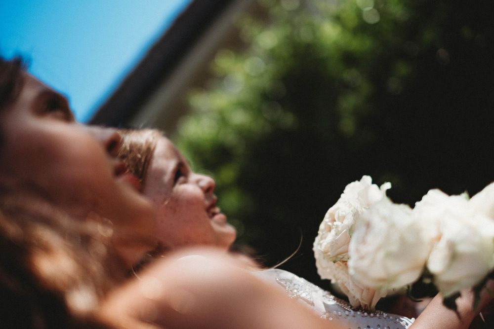 Bride in a sparkling gown laughing joyfully outdoors, holding white roses bouquet on a sunny wedding day.