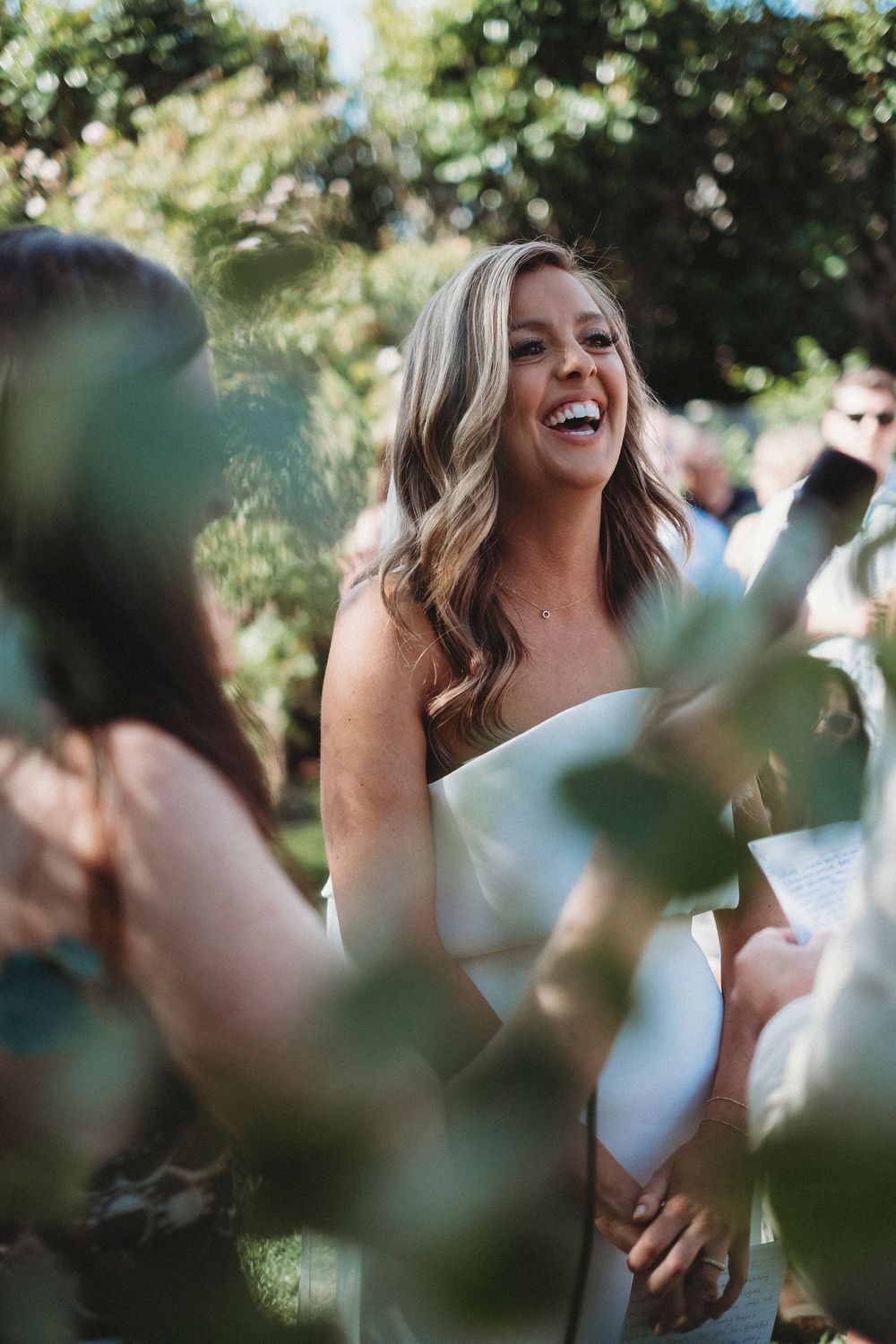 Smiling bride in white strapless gown laughing during outdoor wedding ceremony surrounded by lush green foliage.