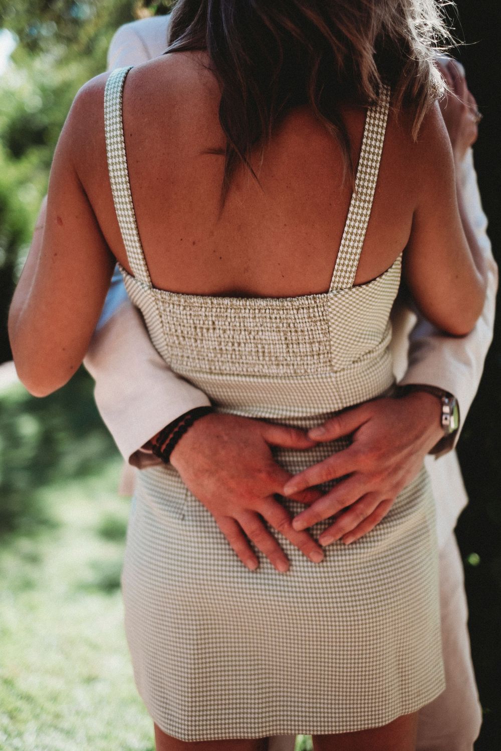 A couple embracing outdoors, mans hands wrapped around womans waist in checkered dress at a wedding.