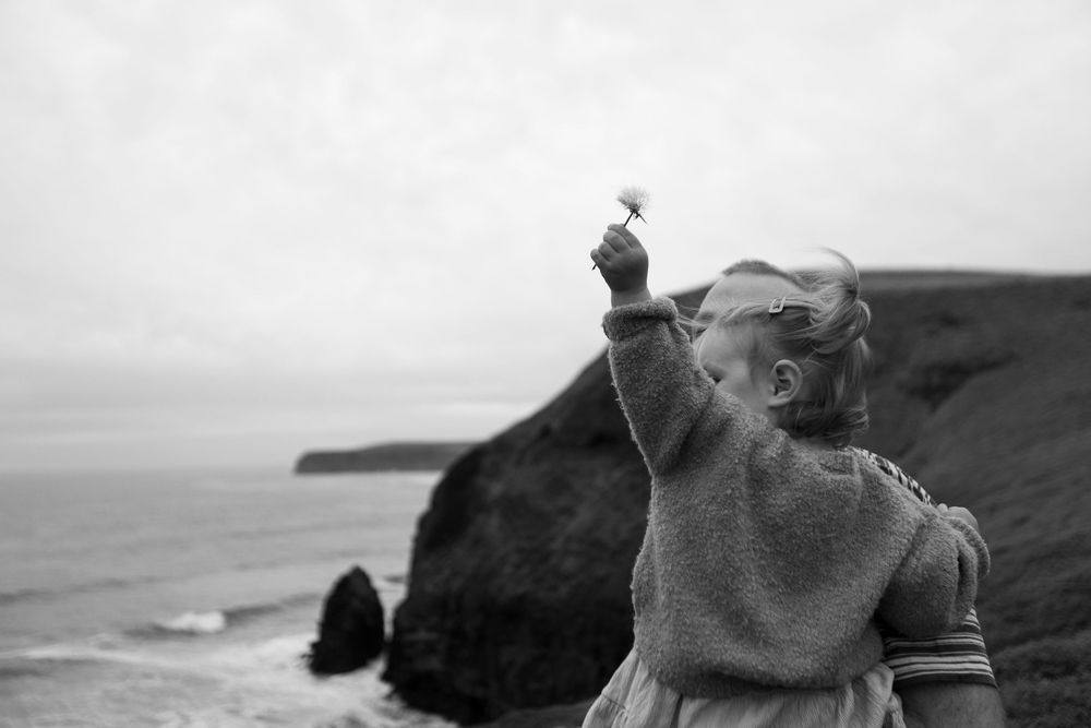 Black and white photo of a young girl holding a flower up toward the sky on a coastal cliff.