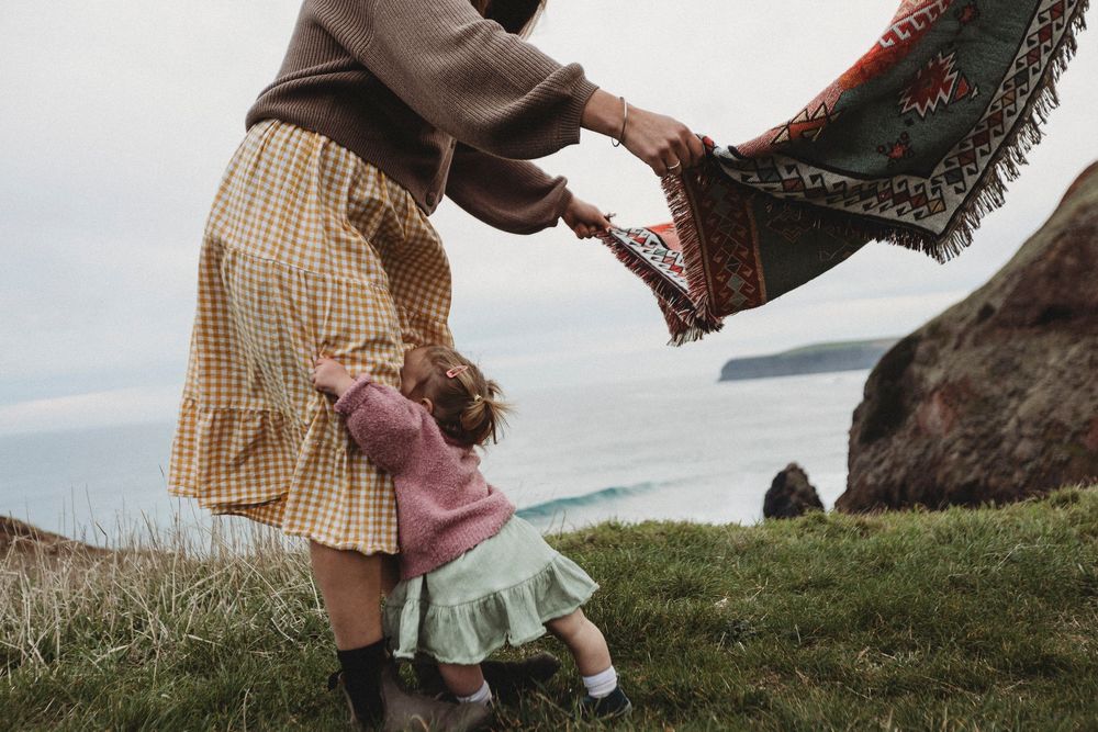 Mother and child playing with a colorful blanket on a coastal cliff with ocean views.